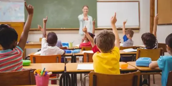 Eine Lehrerin steht vor einer grünen Tafel in einem Klassenzimmer und zeigt auf mehrere Kinder, die an ihren Tischen sitzen und ihre Hände gehoben haben. Die Kinder sind unterschiedlichen Alters und ethnischer Herkunft.