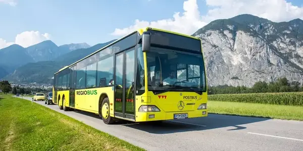 Ein gelber Regiobus der Postbus-Flotte, möglicherweise ein Mercedes-Benz Citaro, fährt auf einer asphaltierten Straße entlang einer grünen Landschaft mit Feldern. Im Hintergrund ragen markante graue Berge unter einem hellen, leicht bewölkten Himmel auf. E