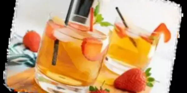 Zwei Gläser mit einem hellbraunen Getränk, Eiswürfeln, Erdbeerscheiben und einem Strohhalm stehen auf einem hölzernen Brett. Frische Erdbeeren liegen daneben. Im Hintergrund ist eine unscharfe Flasche zu erkennen.