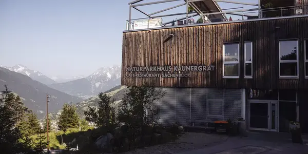 Außenansicht des Naturparkhauses Kaunergrat mit seiner markanten Holzfassade und der Aufschrift "NATURPARKHAUS KAUNERGRAT CAFE-RESTAURANT GACHENBLICK". Im Hintergrund ist eine weite Berglandschaft unter einem hellen Himmel zu sehen.