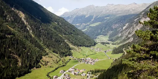 Panoramablick auf ein grünes Tal mit kleinen Ortschaften, umgeben von steilen, bewaldeten Berghängen unter einem teilweise bewölkten Himmel.